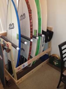 A freestanding DIY surfboard rack made from a 2x4, 1x4, PVC pipe, and pipe insulation is storing 5 surfboards vertically