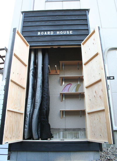 The interior of a wooden surf shed reveals 3 surfboards in board bags and 2 surfboard fins on a storage shelf