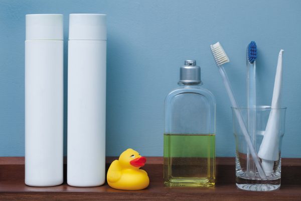 2 white bottles and a rubber ducky, cologne bottle, and glass toothpaste and toothbrush holder on a wooden bathroom shelf 