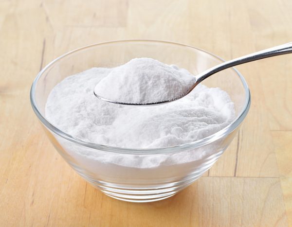 a spoon of baking soda over a glass bowl of baking soda on a wooden table