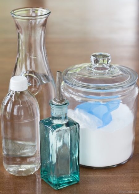 baking soda in a white jar, white vinegar in a clear bottle, a glass water pitcher, and a small blue olive oil bottle 