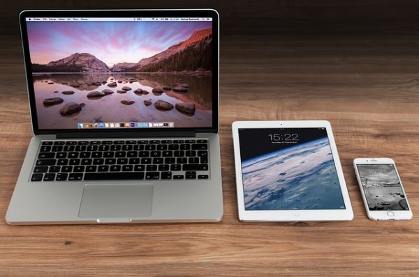 a macbook, white ipad, and white iphone on wooden desk