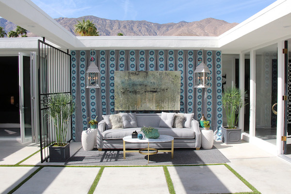 backyard patio with a stylish sofa, coffee table, side tables, a wall-mounted painting, and hanging lanterns, designed by lori dennis