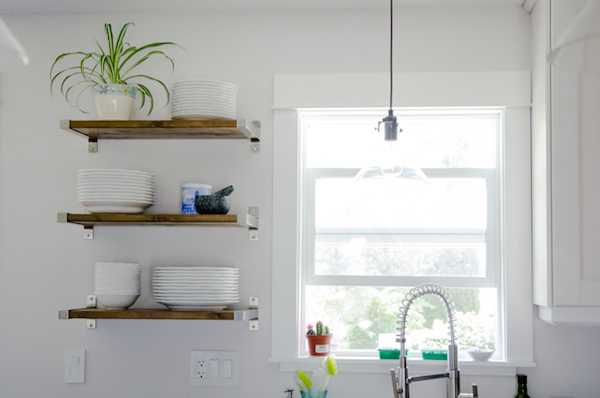 floating shelves hold plates and a plant near a window in a white kitchen 
