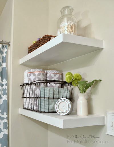 2 white DIY floating shelves storing a wicker tray, glass container of q-tips, wire basket of toiletries, clock, and plant