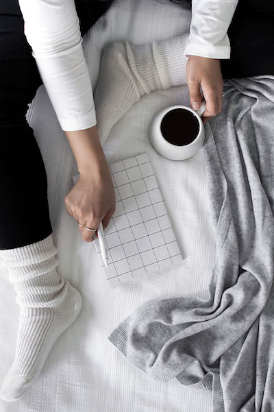 a woman sits on a bed with a mug of tea and a journal