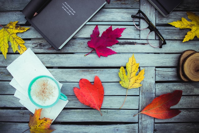 A journal surrounded by a bunch of colorful fall leaves, a pair of reading glasses and a cup of coffee.