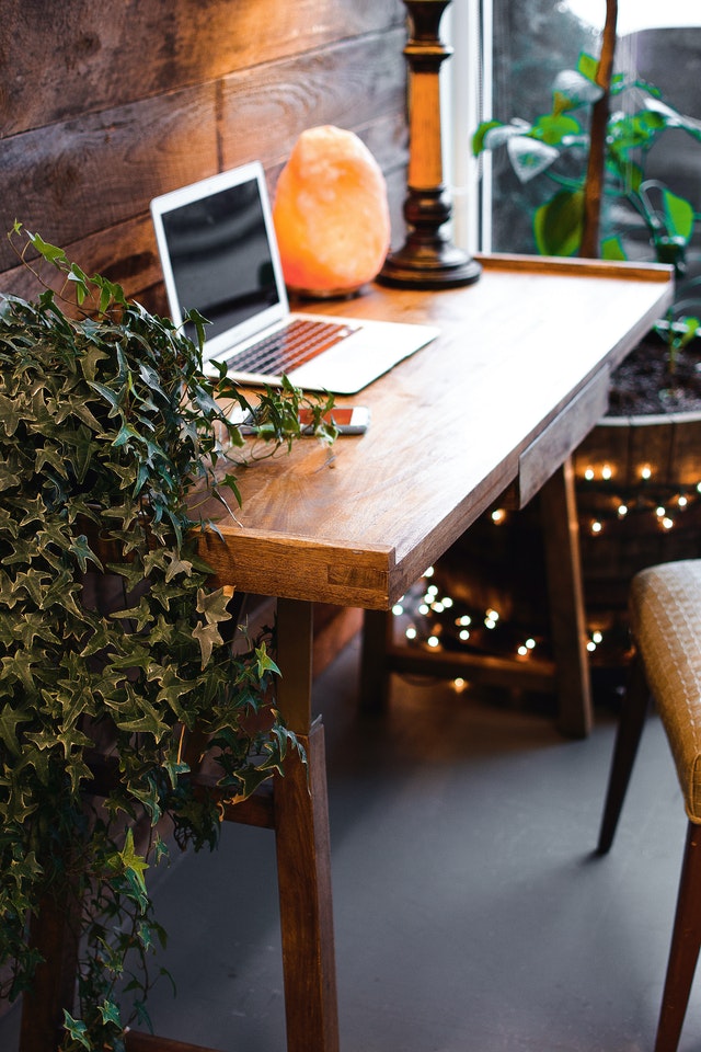 A wooden desk by the window surrounded by plants and a Himalayan salt lamp
