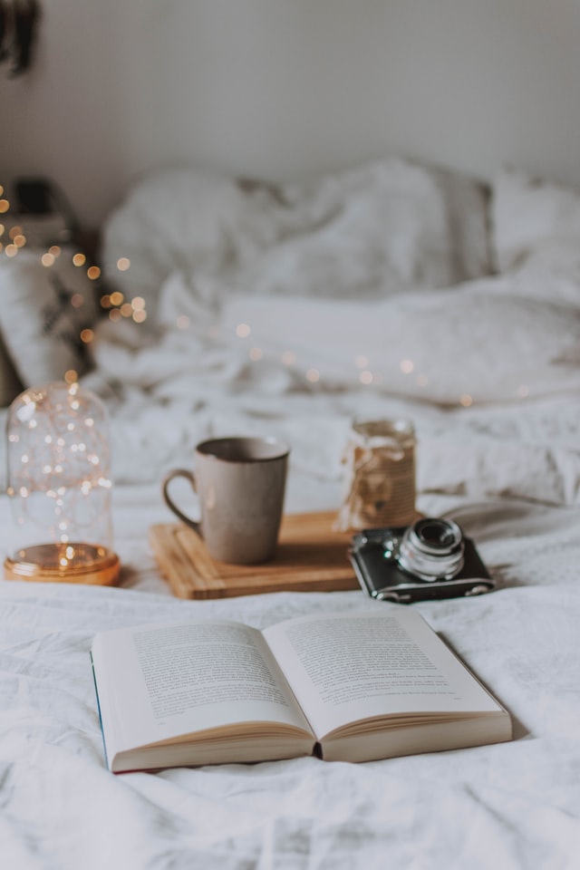 A book laying open on a bed surrounded by fairy lights, a camera and a cup of coffee