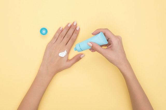 A yellow backdrop and two hands applying a lotion from a blue tube